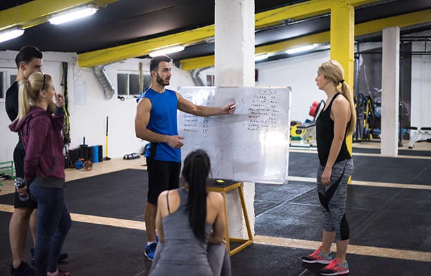 Coach deportivo supervisa a mujer atleta mientras registra resultados en pizarra de gimnasio, ilustrando planificación de proyectos deportivos.