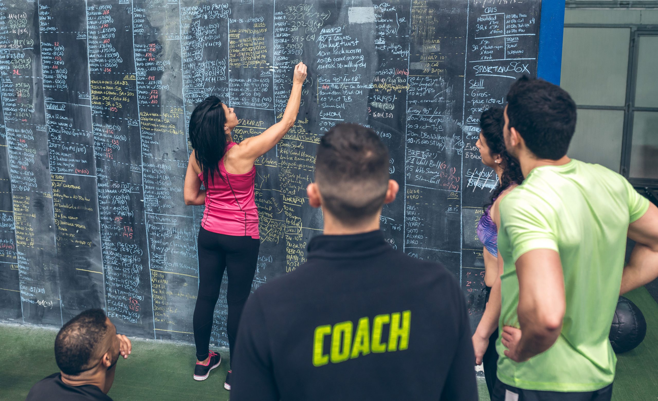 Coach deportivo supervisa a mujer atleta mientras registra resultados en pizarra de gimnasio, ilustrando planificación de proyectos deportivos.