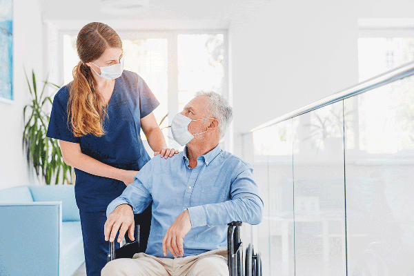 Profesional de enfermería acompaña a persona mayor en silla de ruedas, entregando apoyo cercano y atención humanizada en un entorno de salud.