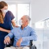 Profesional de enfermería acompaña a persona mayor en silla de ruedas, entregando apoyo cercano y atención humanizada en un entorno de salud.