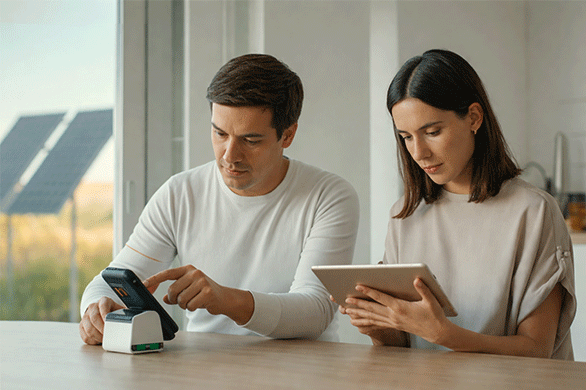 Personas midiendo el consumo eléctrico con medidor digital y tablet para mejorar la eficiencia energética en el hogar con apoyo de energía solar.
