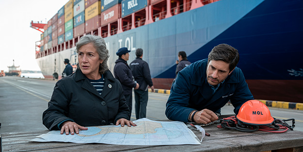 Profesionales marítimos chilenos trabajando en puerto, revisando planos y protocolos de seguridad y reglamentación marítima internacional al lado de un buque. Se observan contenedores sobre el buque y personal marítimo fiscalizando.