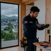Instalador eléctrico trabajando en un tablero de distribución eléctrica en una casa de madera con ventanal panorámico que muestra la bahía y volcanes nevados de Puerto Montt.